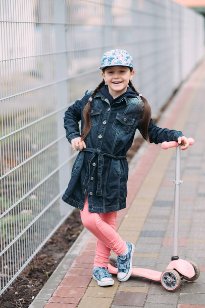 Cute little girl in jeans cardigan and on scooter on the background of the fence - Фото, зображення