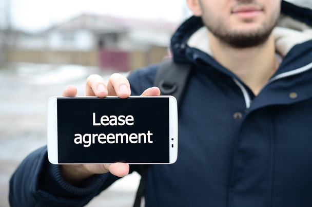 A young guy shows an inscription on the smartphone's display on the background of a big house in winter. Lease agreement - Photo, Image