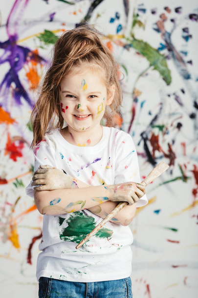 Smiling little girl with painting brush standing in front of messy background. - Foto, Bild