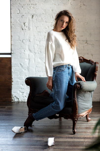 full length view of seductive brunette girl in white sweater and jeans kneeling on armchair and looking at camera - Fotografie, Obrázek