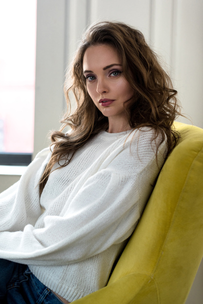 portrait of beautiful tender brunette girl in white jumper sitting on armchair and looking at camera    - Foto, Imagen