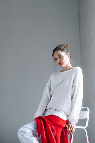 beautiful young woman in trendy white clothes sitting and looking at camera on grey  - 写真・画像