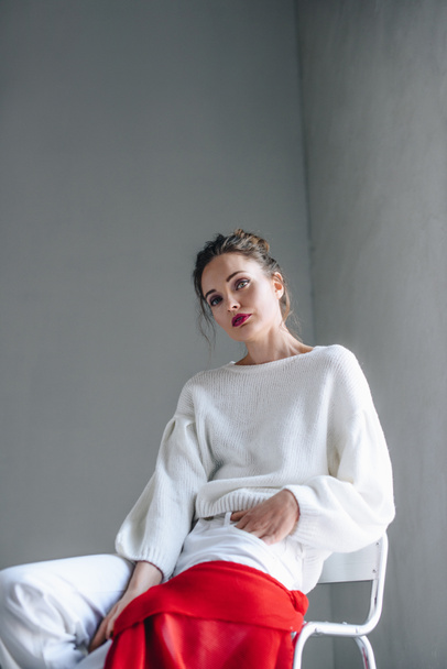 portrait of seductive young woman in trendy white clothes sitting and looking at camera on grey  - Foto, Imagen