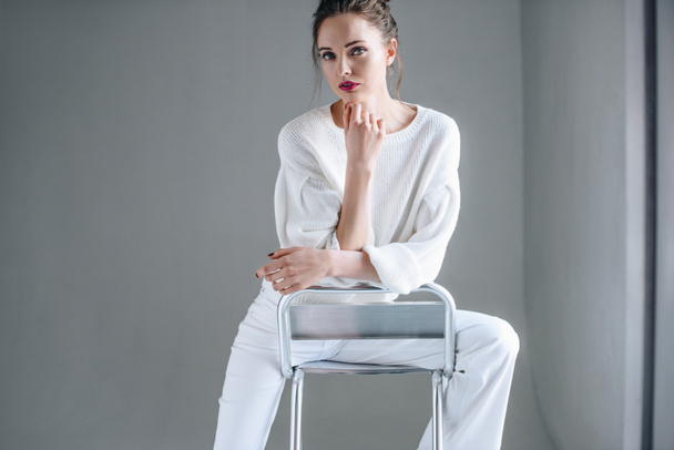 portrait of beautiful brunette woman in trendy white clothes sitting on chair and looking at camera on grey  - Photo, Image