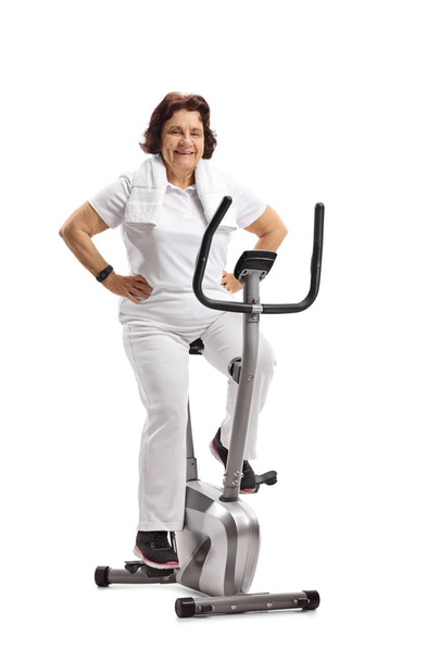Elderly woman sitting on an exercise bike isolated on white background - 写真・画像