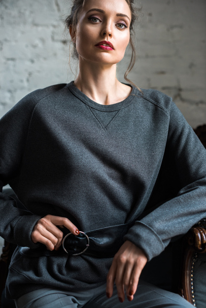 attractive brunette woman in trendy grey jumper with belt sitting on armchair and looking at camera - Foto, afbeelding