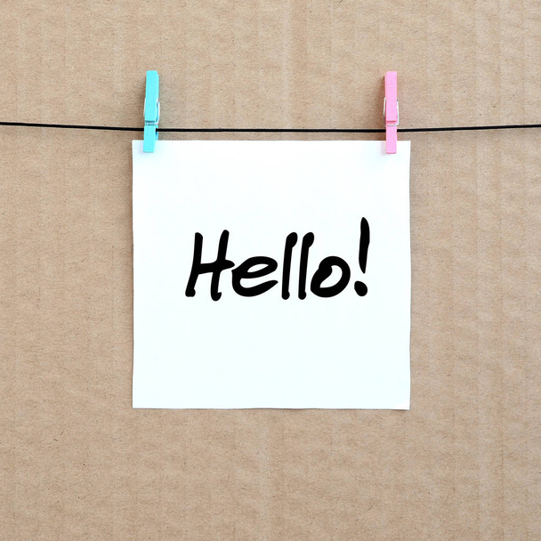 Hello! Note is written on a white sticker that hangs with a clothespin on a rope on a background of brown cardboard - Photo, Image