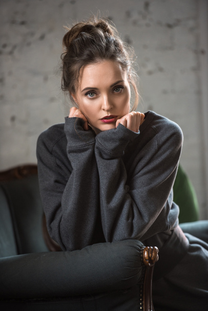 portrait of beautiful brunette woman in trendy sweater sitting on armchair and looking at camera  - Foto, afbeelding