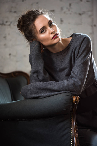portrait of beautiful brunette woman in fashionable clothes sitting on armchair and looking at camera - Фото, изображение