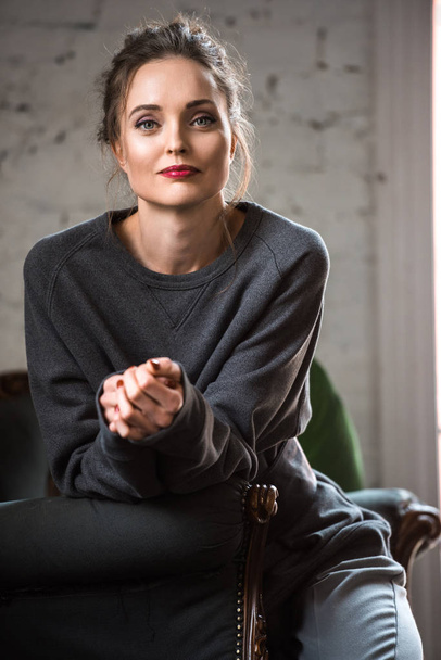 ritratto di attraente donna bruna in maglione grigio seduta sulla poltrona e sorridente alla macchina fotografica
  - Foto, immagini