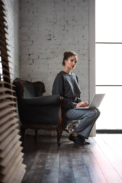full length view of beautiful stylish woman using laptop and looking at camera while sitting on armchair - Fotografie, Obrázek