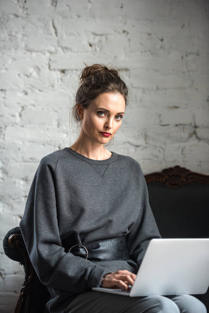 beautiful brunette woman in stylish outfit using laptop and looking at camera - Фото, зображення