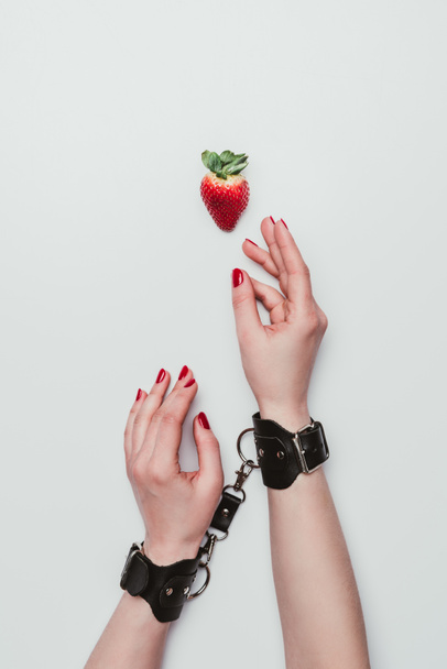 Female hands tied with leather handcuffs reaching for strawberry isolated on white - Photo, image