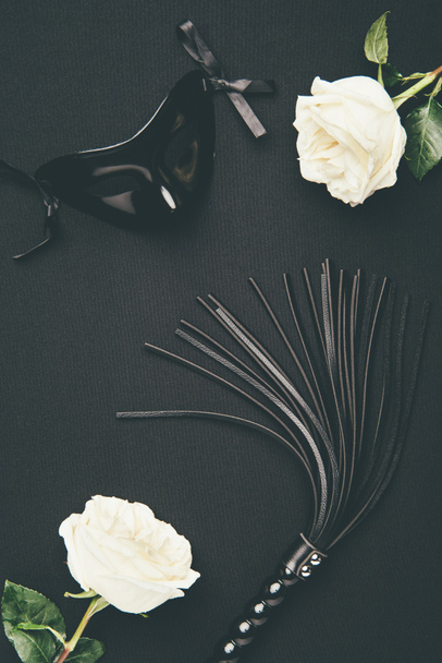 Black flogging whip and mask with white roses isolated on black - Foto, imagen