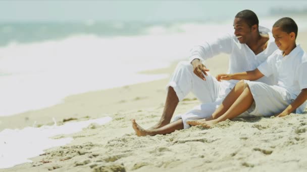 Etnikai boldog apa szórakozás ül fia a strandon - Felvétel, videó