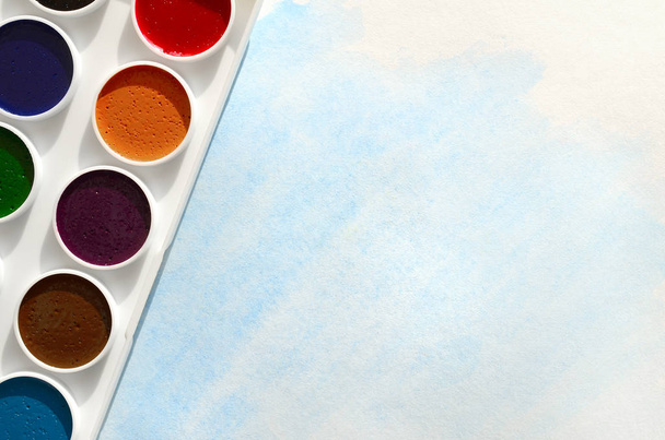 A new set of watercolors lies on a sheet of paper, which shows an abstract watercolor drawing in the form of blue strokes. The concept of amateur painting among teenagers - Photo, Image