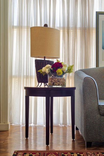 Lampe de table et planteur de fleurs colorées sur petite table en bois brun foncé sur fond de grande fenêtre avec rideaux transparents blancs
 - Photo, image