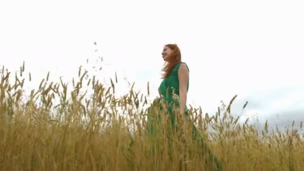 Walk in the mature field.Slow motion. A cheerful red-haired girl in a green dress is walking along the yellow field (oats and wheat).She smiles and laughs. The camera moves next to her. Bottom view. - Footage, Video