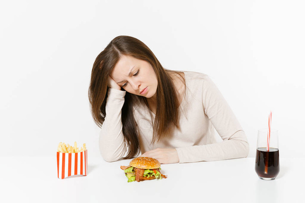 Sad tired woman put hand on head sitting at table with burger, french fries, cola in glass bottle isolated on white background. Proper nutrition or American classic fast food. Area with copy space. - Photo, Image