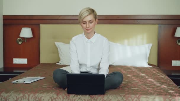 Smiling businesswoman put off laptop computer and taking yoga lotus position sitting relaxed on bed in hotel room. Travel, business and people concept - Video, Çekim