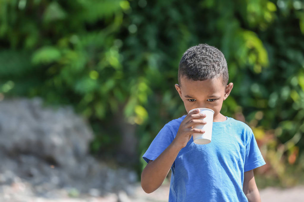 Cute African American boy drinking water from plastic cup, outdoors - 写真・画像