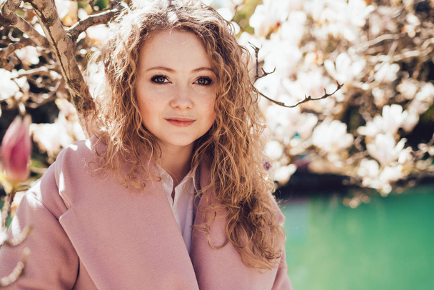 beautiful curly girl in a pink coat enjoying the warm spring weather, walking around the park surrounded by a magnolia and a pond - Photo, image