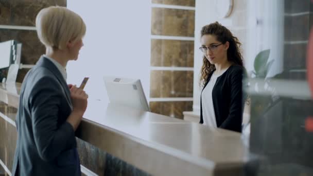 Businesswoman guest in hotel reception paying bill contactless payment credit card technology and talking with receptionist girl standing at desk. Business, travel and people concept - Footage, Video