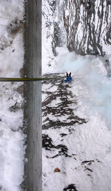 青いジャケットの懸垂下降で男性アイスクライマー、垂直の凍った滝に橋の欄干から - 写真・画像