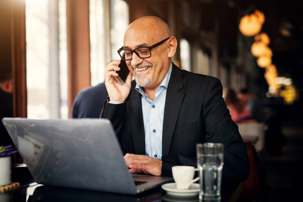 mature businessman using laptop, talking over mobile phone and drinking coffee in cafe  - Photo, Image