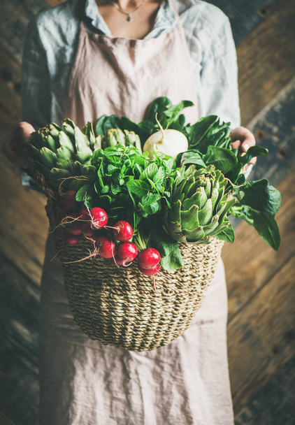 Female farmer in linen apron holding basket of fresh garden vegetables and greens in her hands - 写真・画像