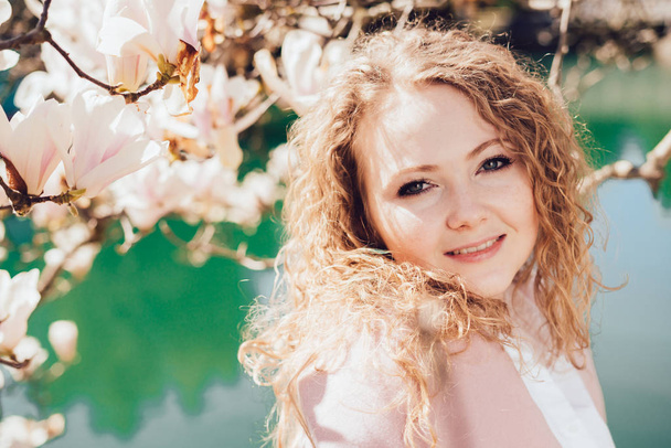 cute curly-haired girl posing in the park in the rays of the spring sun, enjoying magnolia and a pond - Fotoğraf, Görsel