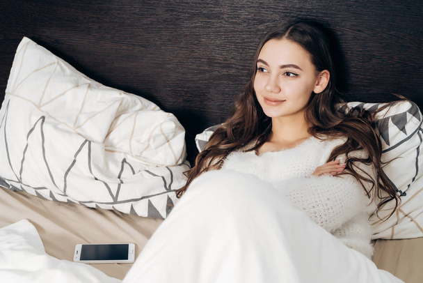 long-haired young girl sits in bed under a white blanket, enjoys a good morning and a day off - 写真・画像