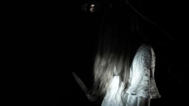 Una niña fantasma con el pelo largo y negro, en blanco, vagando por el bosque con un cuchillo y un juguete suave. 4k
 - Metraje, vídeo