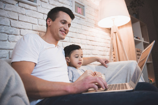 Father and son watching movie on laptop on couch at home - 写真・画像