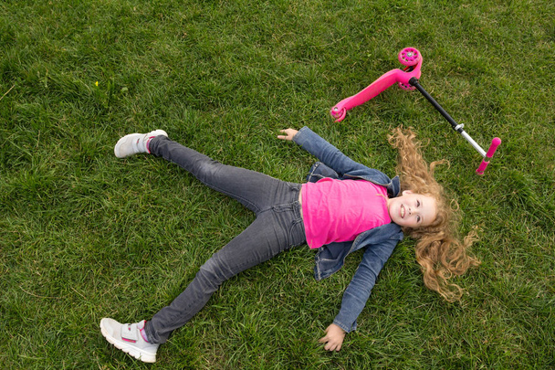 Glimlachend kind meisje dragen jeans jas liggen op gras met roze scooter - Foto, afbeelding