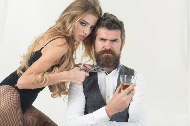 Bearded man drink whiskey, sexy woman with long curly hair. Woman with razor, comb cut hair of man. Couple make haircut, love relations. Barber, hipster fashion, beauty. Couple in love at hairdresser. - Photo, image