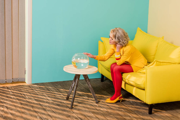 mujer en ropa retro brillante mirando a los peces del acuario mientras descansa en el sofá en el apartamento colorido, concepto de casa de muñecas
 - Foto, imagen