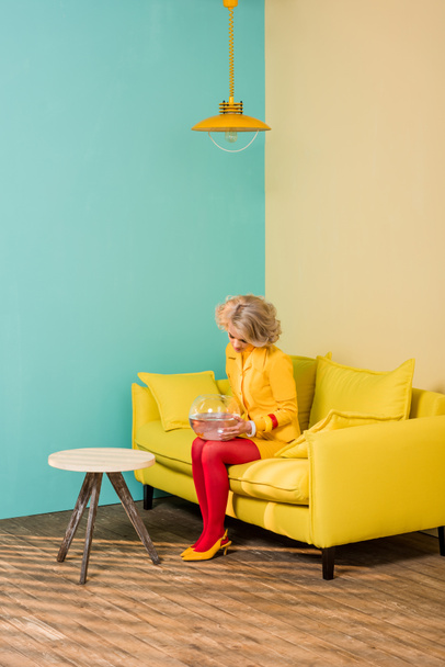 woman in bright retro clothing with golden fish in aquarium resting on sofa at colorful apartment, doll house concept - Photo, Image
