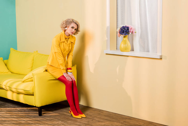 pensive woman in retro clothing sitting on yellow sofa at colorful apartment, doll house concept - Foto, afbeelding