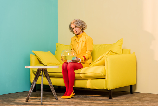 woman in bright retro clothing with golden fish in aquarium on sofa at colorful apartment, doll house concept - Fotó, kép