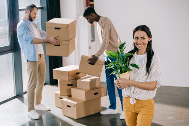 junge Frau hält Topfpflanze in der Hand und lächelt in die Kamera, während männliche Kollegen beim Umzug Schachteln hinter sich halten - Foto, Bild