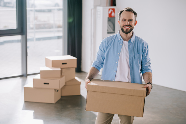 happy bearded businessman holding cardboard box and smiling at camera while moving in new office - Fotografie, Obrázek
