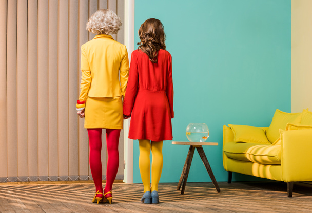 back view of women in bright retro styled clothing holding hands at colorful apartment, doll house concept - 写真・画像