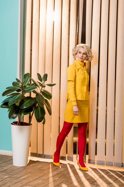 pensive retro styled woman standing at ficus plant in flowerpot at colorful apartment, doll house concept - Photo, Image