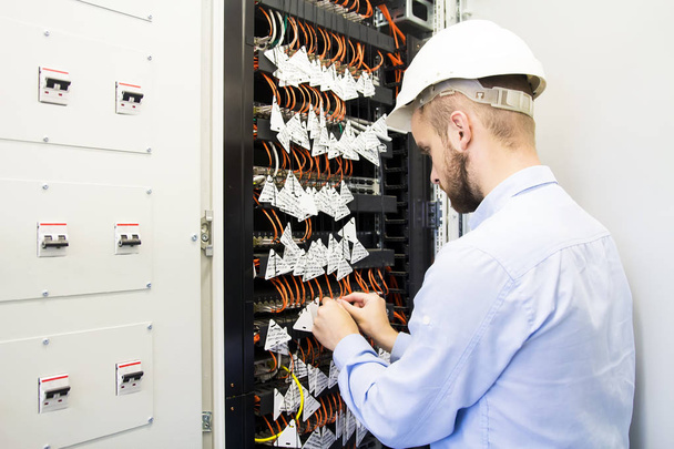 Engineer performs adjustment of computer network cabinet with controllers, switches and optical cables. A worker in white helmet connects - Fotografie, Obrázek