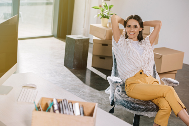 счастливая молодая женщина сидит с руками за головой и улыбается перед камерой в новом офисе
 - Фото, изображение