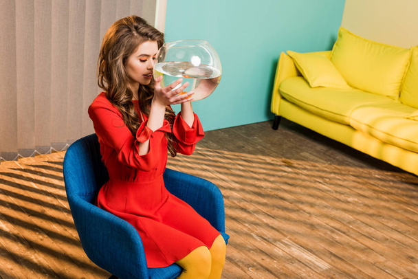 fiatal nő, retro ruházat, aranyhal akvárium ül a szék színes apartman, baba house koncepció - Fotó, kép
