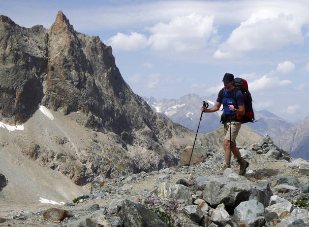 male athletic hiker on his way to a high alpine base camp for mountain climbing walks along a dusty hiking trail in the Barre des Ecrins National Park in the French Alps carrying a large backpack - Photo, Image