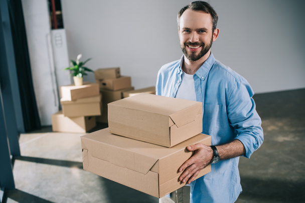 happy bearded man holding boxes and smiling at camera during relocation - Fotoğraf, Görsel