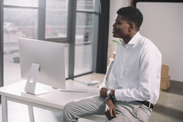 joven y pensativo empresario afroamericano sentado en la mesa y mirando hacia otro lado en una nueva oficina
 - Foto, Imagen